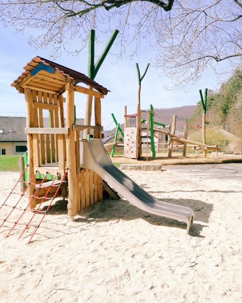 Spielplatz an den Siebenquellen in Geislingen mit Klettergerüst, Rutsche und Sandfläche aus Holz und naturnahen Materialien unter Bäumen