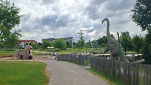 Blick auf den Himmelsgarten in Schwäbisch Gmünd mit großem Spielgelände, Einhorn-Spielgerät, Dinosaurierfigur, Wegen, Grünflächen und bewölktem Himmel.