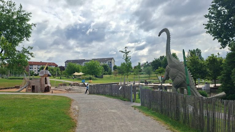 Blick auf den Himmelsgarten in Schwäbisch Gmünd mit großem Spielgelände, Einhorn-Spielgerät, Dinosaurierfigur, Wegen, Grünflächen und bewölktem Himmel.