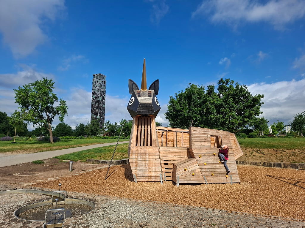 Hölzernes Einhorn-Spielgerät im Himmelsgarten Schwäbisch Gmünd mit Kletterelementen, Grünflächen und Aussichtsturm im Hintergrund.