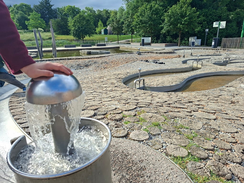 Wasserspielplatz mit sprudelnder Wasserpumpe, geschwungenen Wasserläufen und grüner Parklandschaft im Hintergrund.
