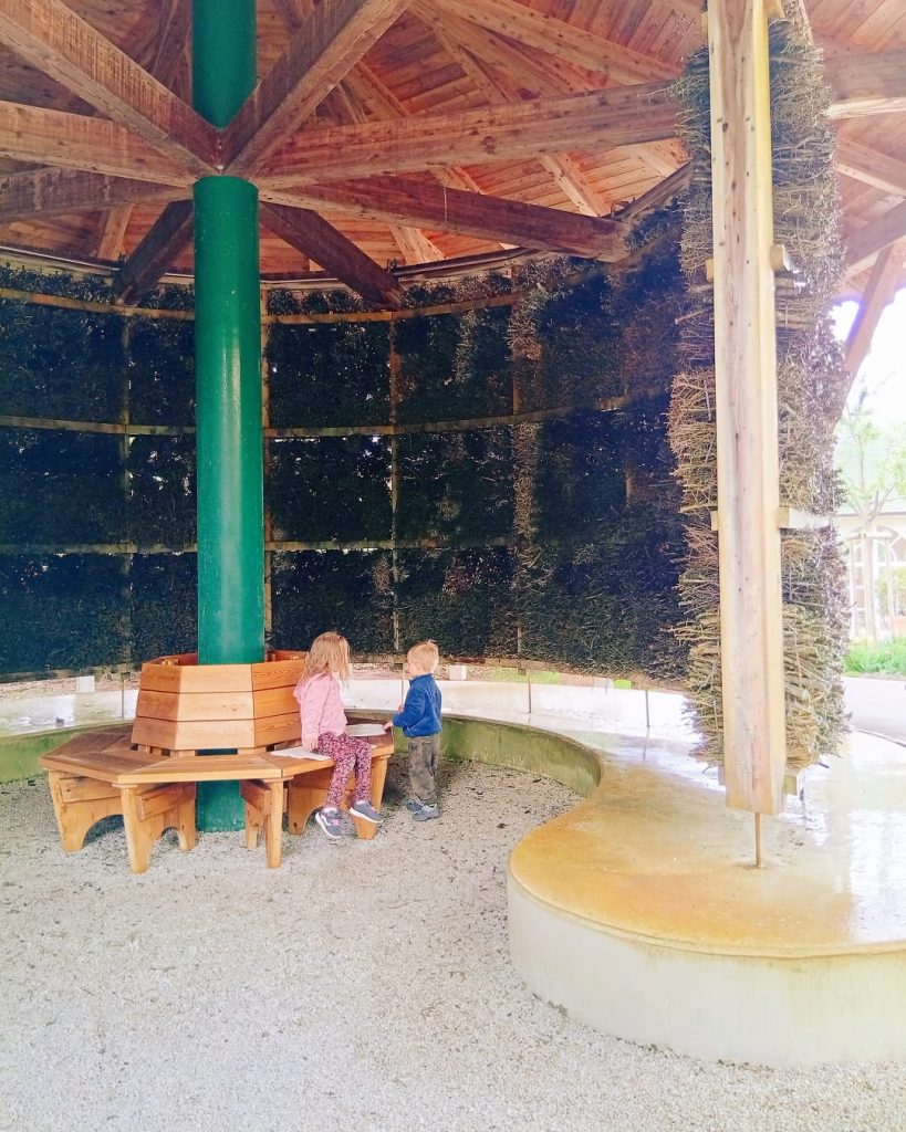 Zwei Kinder sitzen und stehen in einem hölzernen Meeresluft-Inhalatorium mit salzhaltigen Reisigwänden und rundem Wasserbecken.