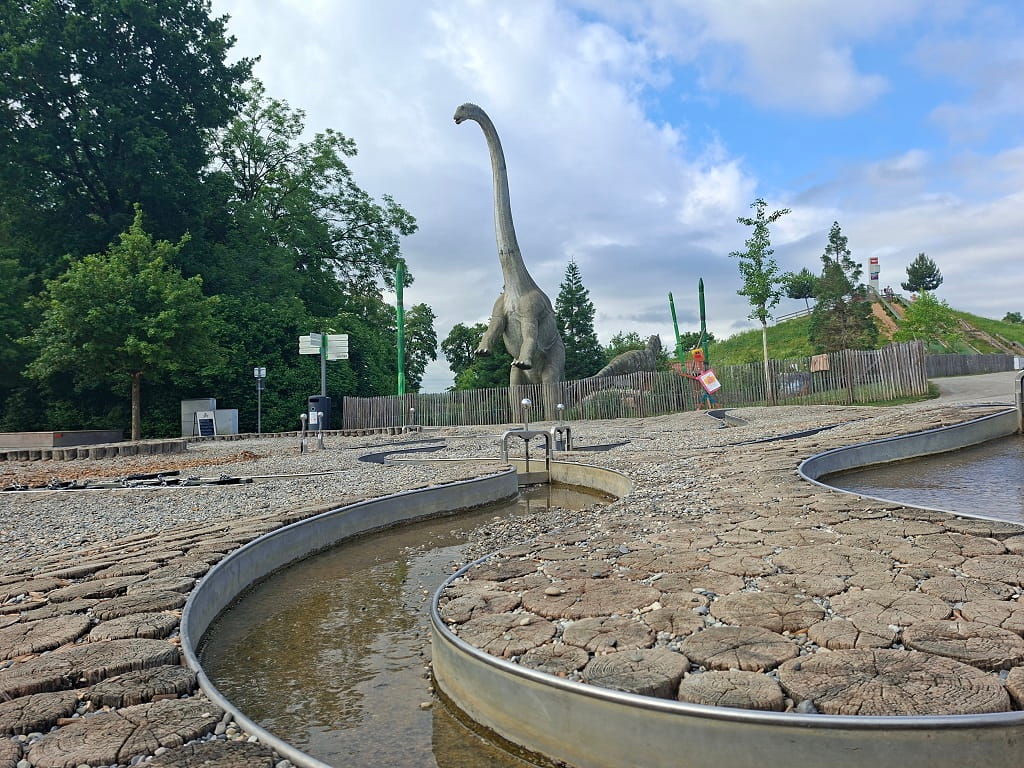 Wasserspielplatz mit geschwungenen Wasserläufen, Dinosaurierfiguren und Grünflächen im Hintergrund.