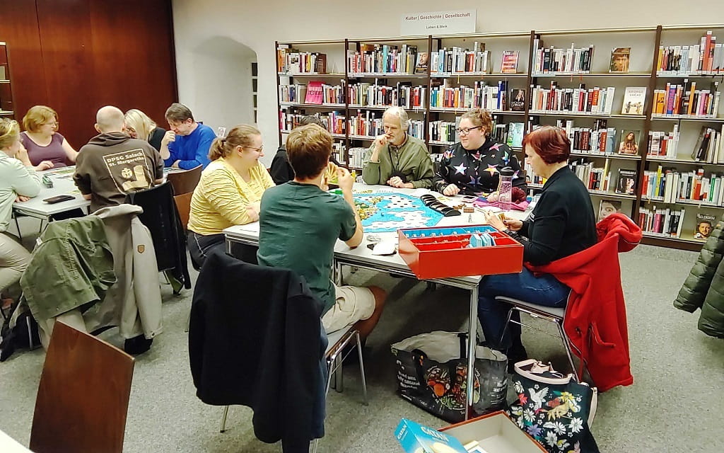 Mehrere Personen sitzen in der Stadtbibliothek Göppingen an zwei Tischen und spielen gemeinsam Brettspiele. Im Hintergrund sind Bücherregale zu sehen, auf den Tischen liegen Spielpläne, Karten und Spielmaterial.