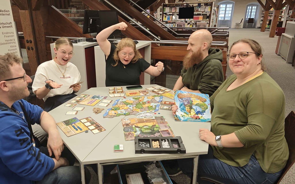 Fünf Personen sitzen in der Stadtbibliothek Göppingen an einem Tisch und spielen gemeinsam ein Brettspiel. Eine Spielerin jubelt mit erhobenen Armen, während die anderen lächeln und Spielkarten, Spielplan und Figuren auf dem Tisch liegen.