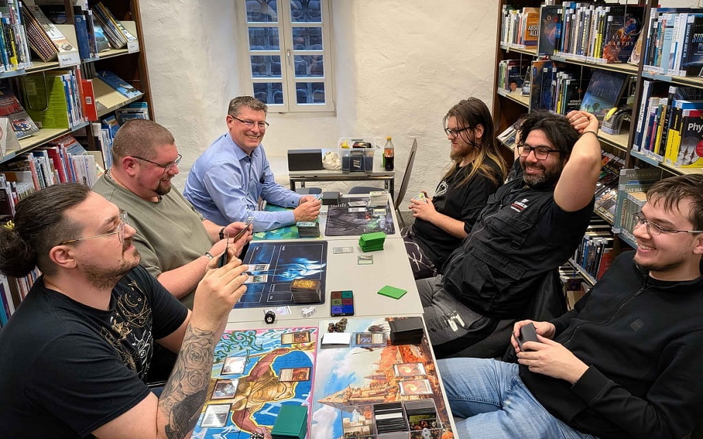 Sechs Personen sitzen in der Stadtbibliothek Göppingen an einem Tisch und spielen gemeinsam ein Kartenspiel. Auf dem Tisch liegen Spielmatten, Kartenstapel, Würfel und Spielmaterial, im Hintergrund sind Bücherregale zu sehen.