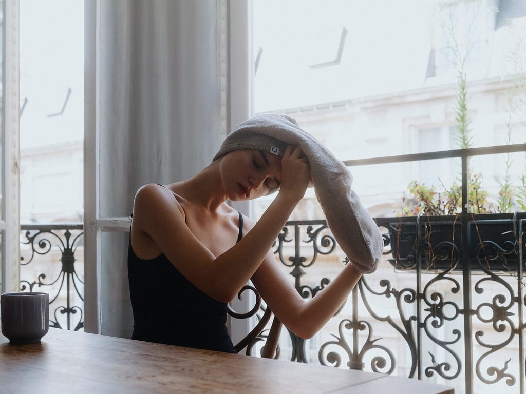 Frau sitzt mit grauem Turban-Handtuch am Fenster.