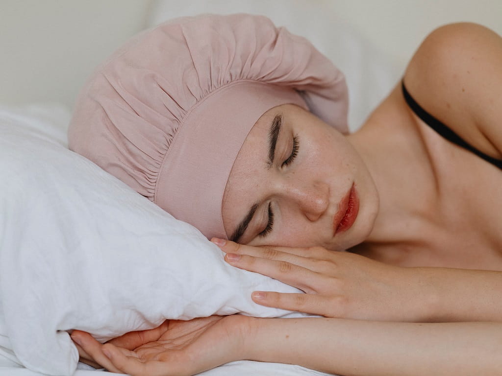 Frau schläft mit rosa Bonnet auf einem weißen Kissen und schützt ihr Haar über Nacht vor Reibung.