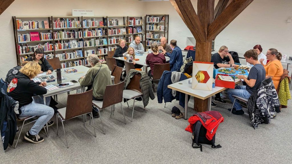 Mehrere Spielrunden des FRoG e. V. sitzen in der Stadtbibliothek Göppingen an verschiedenen Tischen. Einige Personen spielen Brettspiele, andere unterhalten sich oder bereiten Spielmaterial vor. Im Hintergrund sind Bücherregale und der Themenbereich „Kultur | Geschichte | Gesellschaft“ zu sehen.