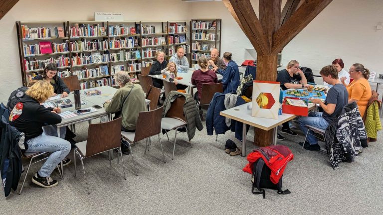 Mehrere Spielrunden des FRoG e. V. sitzen in der Stadtbibliothek Göppingen an verschiedenen Tischen. Einige Personen spielen Brettspiele, andere unterhalten sich oder bereiten Spielmaterial vor. Im Hintergrund sind Bücherregale und der Themenbereich „Kultur | Geschichte | Gesellschaft“ zu sehen.