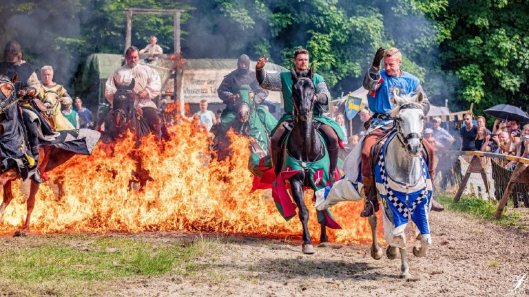 Mehrere Ritter reiten beim StauferSpektakel vor einer Feuerwand über den Turnierplatz, während das Publikum im Hintergrund zuschaut.