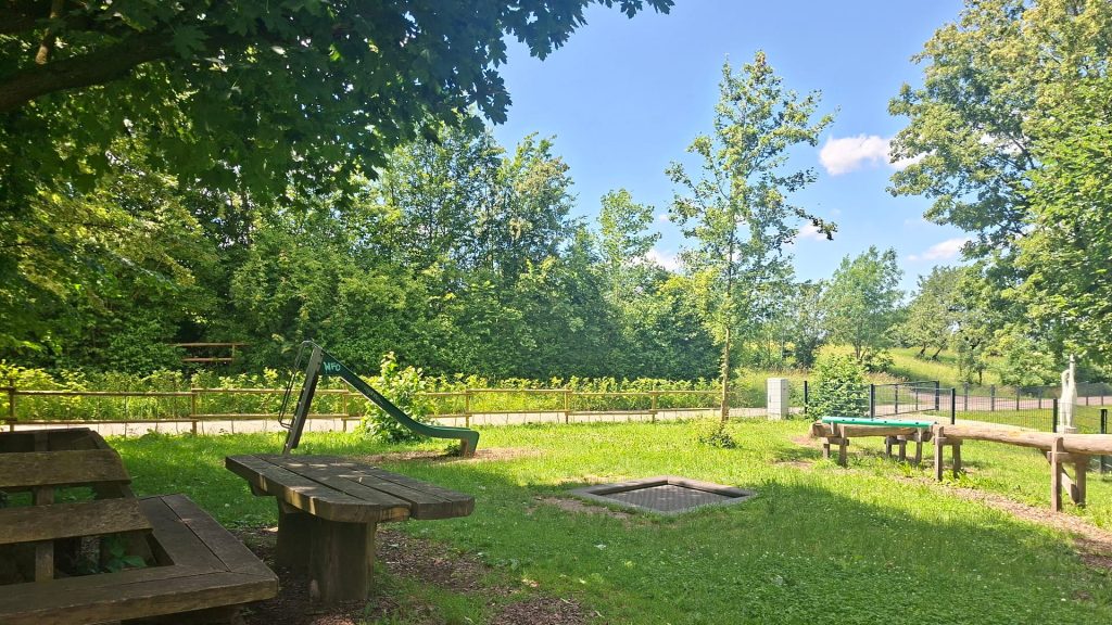 Spielplatz am Feuersee in Dürnau mit Rutsche, Trampolin, Sitzbänken und Tischtennisplatte auf einer grünen Wiese unter Bäumen.
