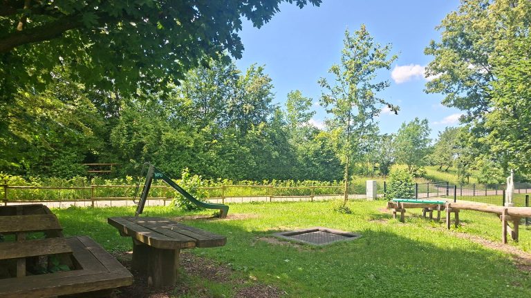 Spielplatz am Feuersee in Dürnau mit Rutsche, Trampolin, Sitzbänken und Tischtennisplatte auf einer grünen Wiese unter Bäumen.