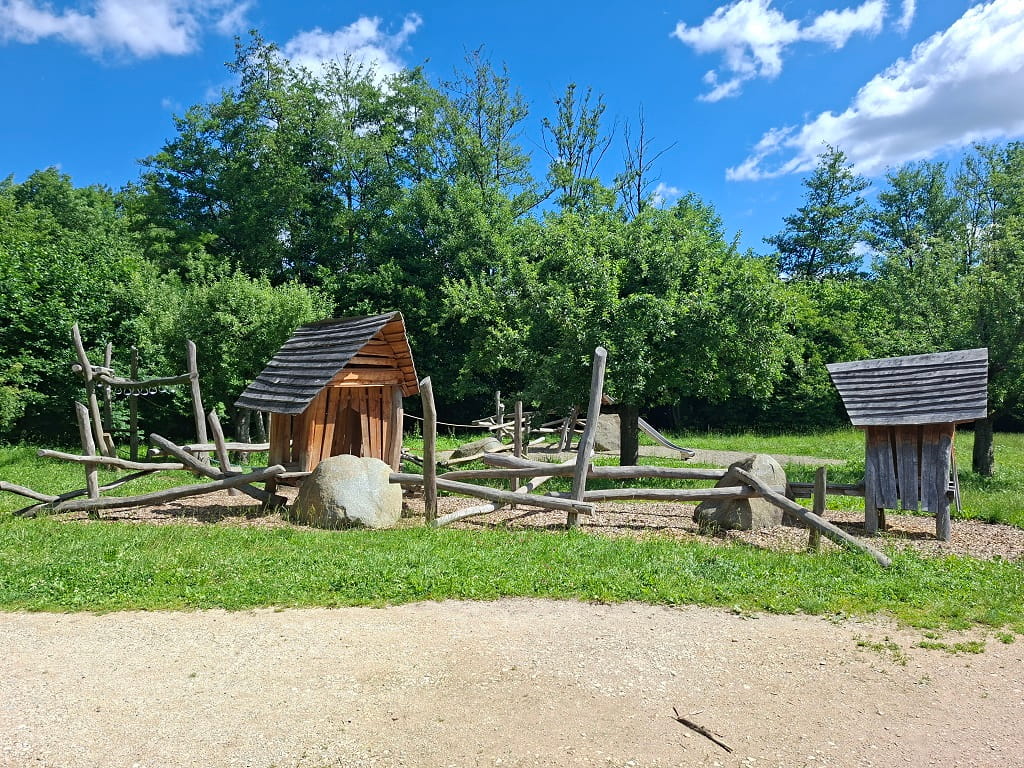 Idyllischer Naturspielplatz mit Holzhütten, Kletterelementen und grüner Umgebung.