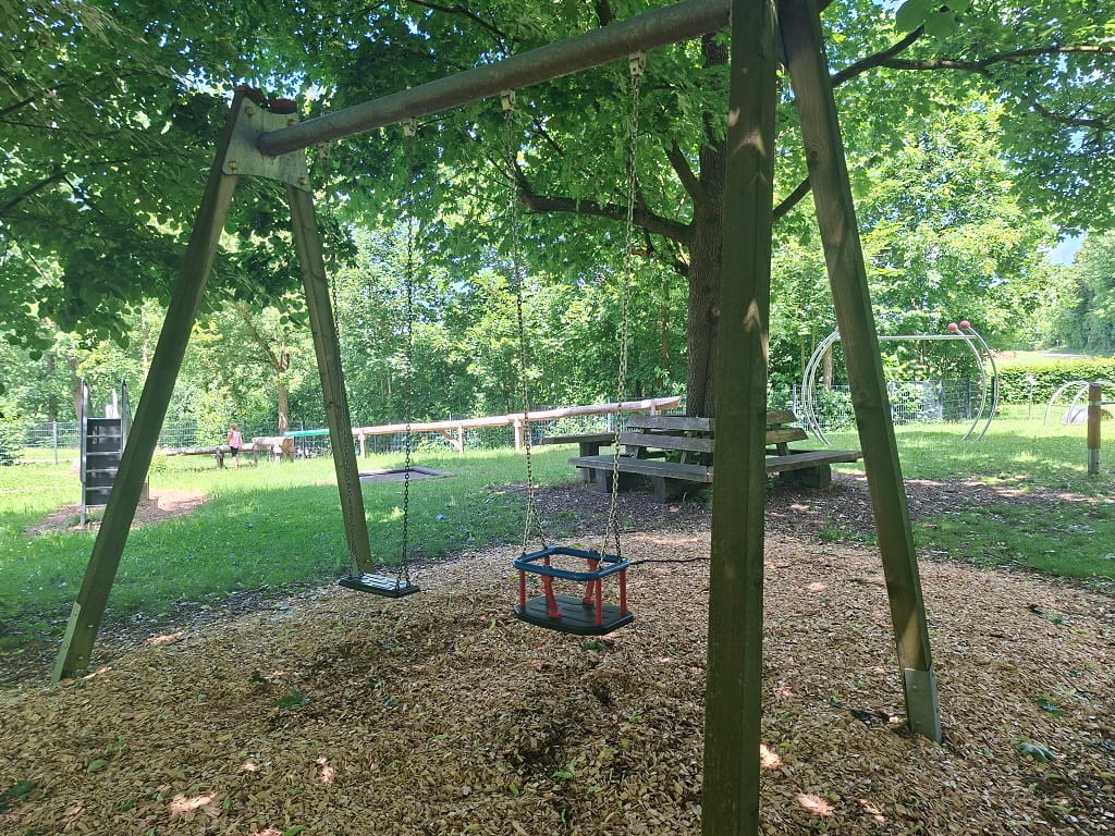 Schaukelanlage mit Brettschaukel und Kleinkindschaukel auf dem Spielplatz am Feuersee in Dürnau, umgeben von Bäumen und Sitzbänken.