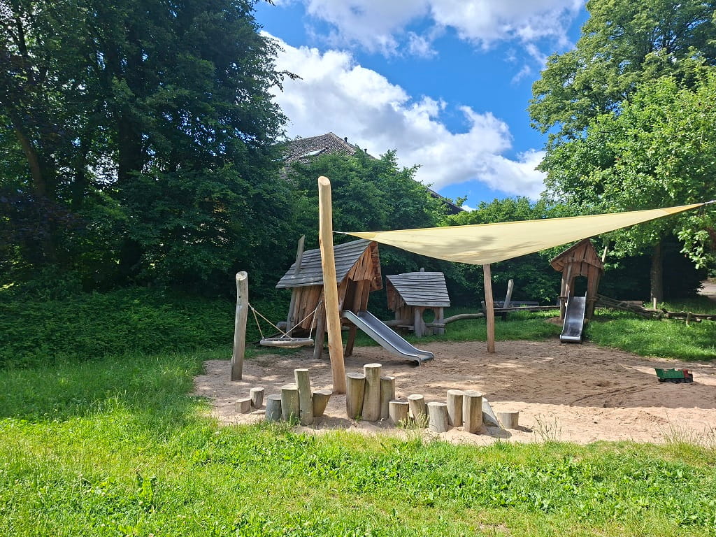 Spielbereich mit Rutschen, Holzhütten, Sandfläche und Sonnensegel.
