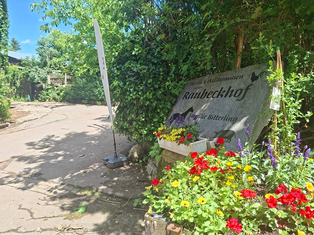 Eingangsbereich des Raubeckhofs in Dürnau mit beschriftetem Steinschild, Blumenbeeten und viel Grün am Wegesrand.