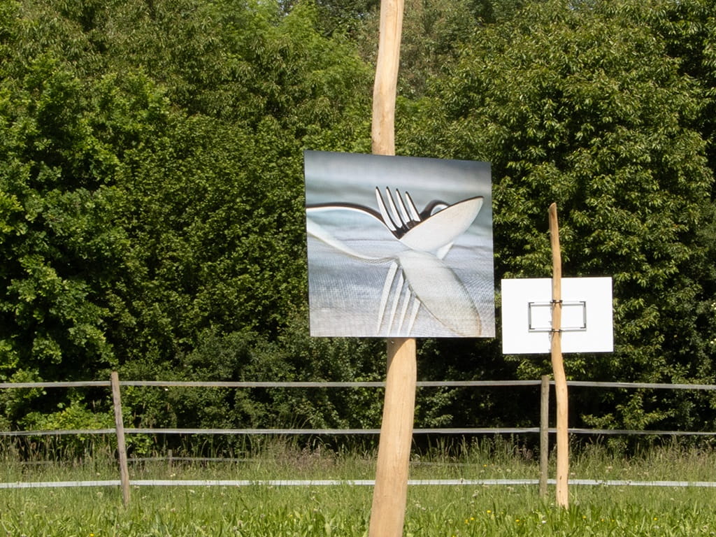 Eine Fotografie mit Messer und Gabel hängt auf einer Holzstele im Grünen und zeigt die Motivvielfalt der Jubiläumsausstellung des Fotoclubs Albershausen.