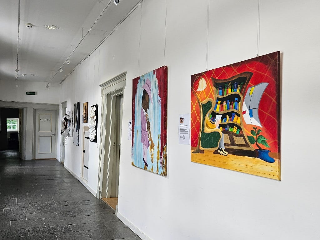 Blick in die Michael-Ferk-Ausstellung im Nordflügel von Schloss Filseck mit mehreren farbigen Werken an einer weißen Wand, darunter das Bild „Das Wohnzimmer“.
