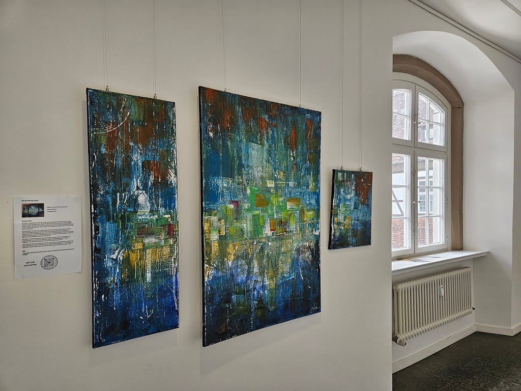Drei abstrakte Bilder des Werks „Die Leuchtende Stadt“ von Michael Ferk hängen in einer Ausstellung auf Schloss Filseck neben einem Fenster.