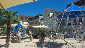 Kinder und Familien auf dem Pop-up-Spielplatz in Schwäbisch Gmünd mit großem Klettergerüst, Sandfläche und Palmen zwischen den Gebäuden der Altstadt.