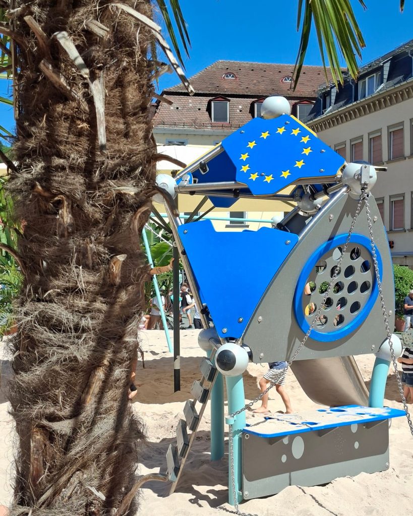 Blaues Kletter- und Spielgerät mit Europa-Sternen auf dem Pop-up-Spielplatz in Schwäbisch Gmünd, eingerahmt von einer Palme und historischen Gebäuden.