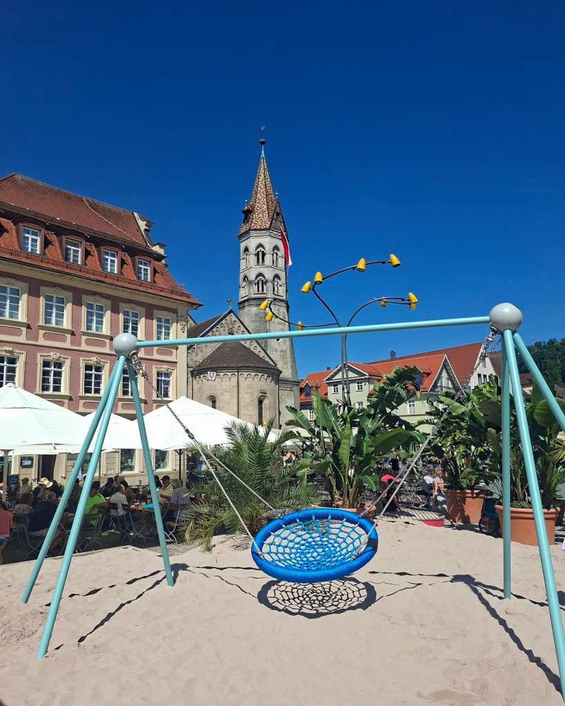 Blaue Nestschaukel im Sand auf dem Pop-up-Spielplatz in Schwäbisch Gmünd, mit Palmen, Außengastronomie und dem Johannisturm im Hintergrund.