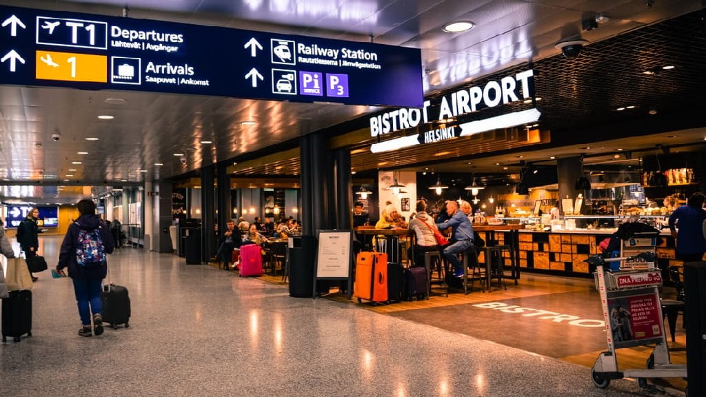 Bistrobereich in einem Flughafen mit Reisenden, Koffern und Hinweisschildern zu Abflug und Ankunft.