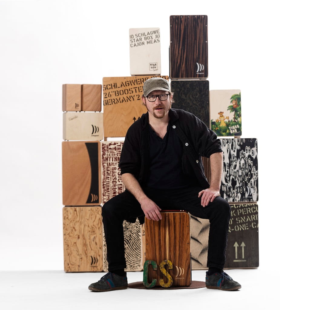 Martin „Mädde“ Sauer, Cajonlehrer und Berufsmusiker, sitzt vor mehreren Cajons für den Artikel zum Cajon-Workshop auf dem Hohenstaufen.