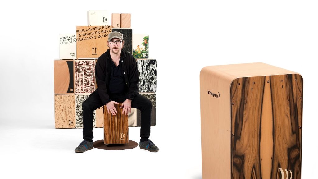 Martin „Mädde“ Sauer sitzt auf einer Cajon vor mehreren Cajons neben einer Schlagwerk-Cajon für den Cajon-Workshop auf dem Hohenstaufen.
