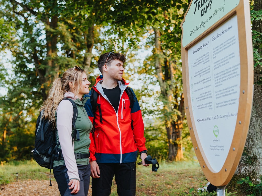 Zwei Wandernde mit Rucksäcken lesen die Informationstafel des Trekking-Camps im Wald bei Gingen an der Fils.