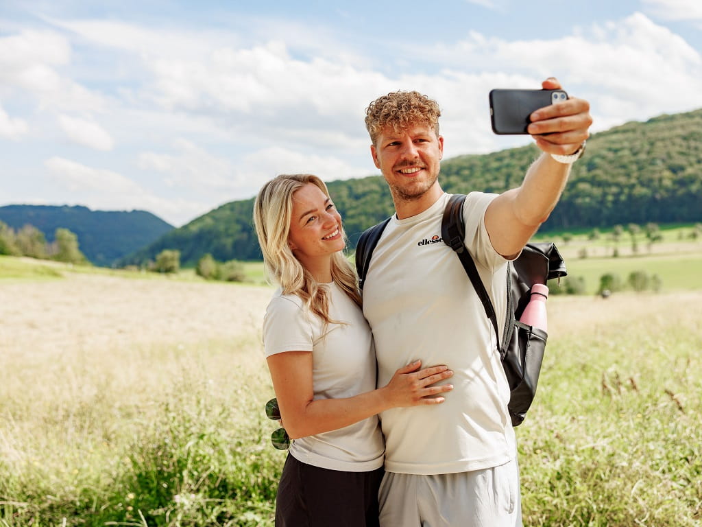 Paar macht ein Selfie vor einer hügeligen Landschaft.