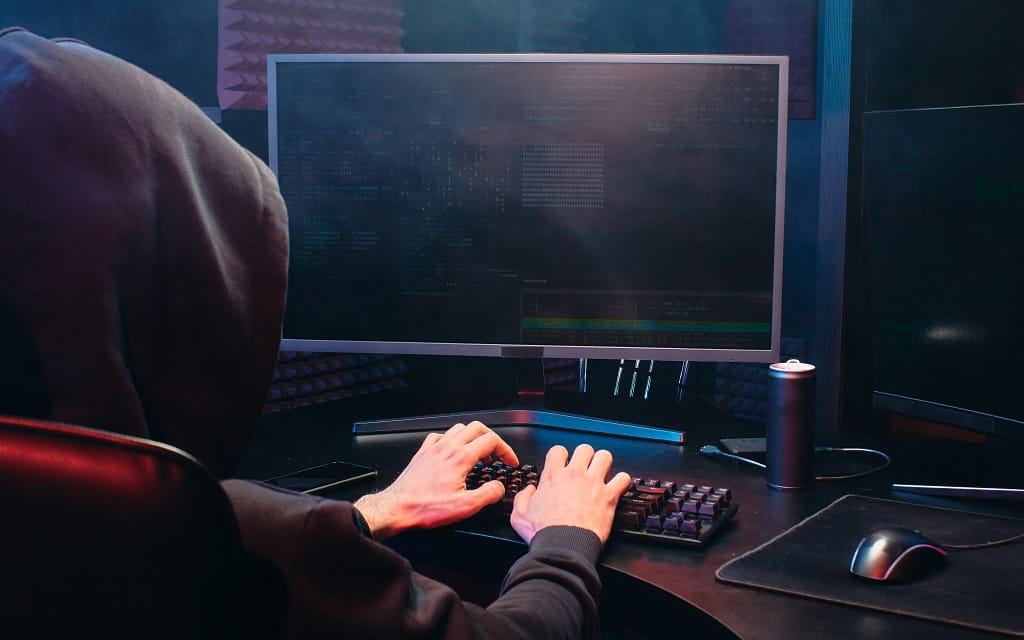 Person mit schwarzem Kapuzenpullover sitzt vor mehreren Computermonitoren und tippt auf einer Tastatur.