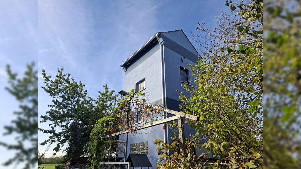 Blauer Bücherturm in Dürnau mit begrüntem Außenbereich, Holzpergola und bunten Dekorationen vor strahlend blauem Himmel.