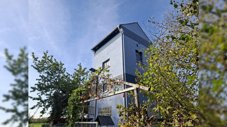 Blauer Bücherturm in Dürnau mit begrüntem Außenbereich, Holzpergola und bunten Dekorationen vor strahlend blauem Himmel.