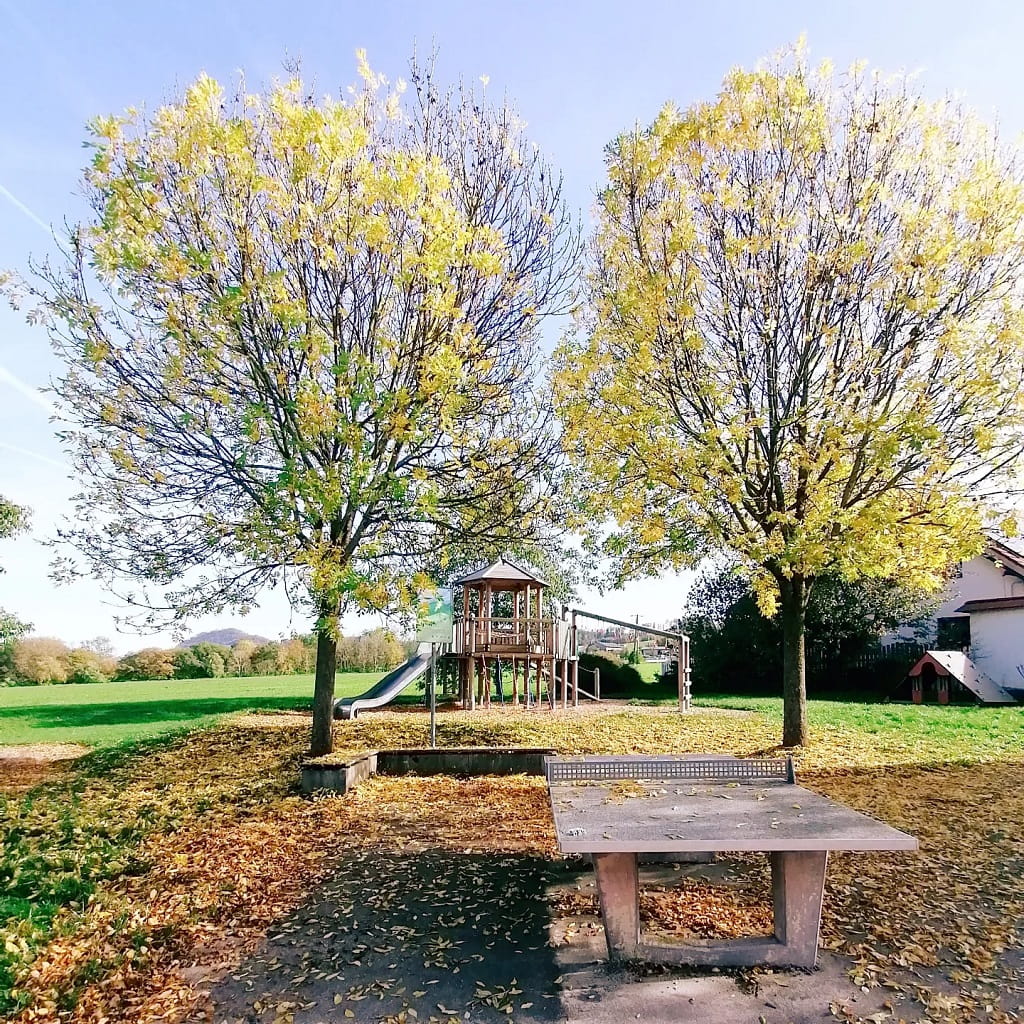 Spielplatz in Göppingen-Maitis mit einem Spielturm samt Rutsche und einer Tischtennisplatte zwischen zwei großen Bäumen.