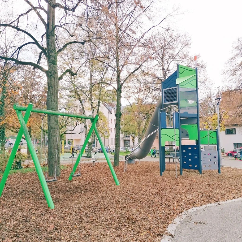 Spielplatz am Erich-Mühsam-Platz in Göppingen-Holzheim mit einem hohen Kletterturm samt Röhrenrutsche und einer Doppelschaukel unter großen Bäumen.