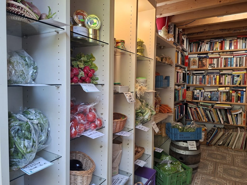 Regale mit frischem Gemüse, Radieschen, Tomaten, Salat und weiteren Lebensmitteln neben Bücherregalen im blauen Bücherturm in Dürnau.